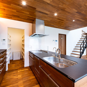 木のぬくもりを大切にした次世代住宅 従来のキッチンとは一線を画す、デザイン性のあるアイランドキッチン。優しい風合いが素敵なブラックウォールナット天然木突板扉に硬質なセラミックのワークトップと素材感抜群。スタイリッシュな家具として存在感のあるしつらえとなっています。ダイニングキッチンの天井には床材と同じチークを使用しており、建具などのトーンも合わせることで重厚感のある空間に仕上がです。間接照明を施すことで空間のアクセントとなっています。 福岡工務店 福岡注文住宅建設 福岡注文住宅